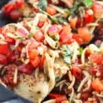 A close-up of a skillet with meat topped with a mixture of diced tomatoes, shredded cheese, red onions, and fresh basil.