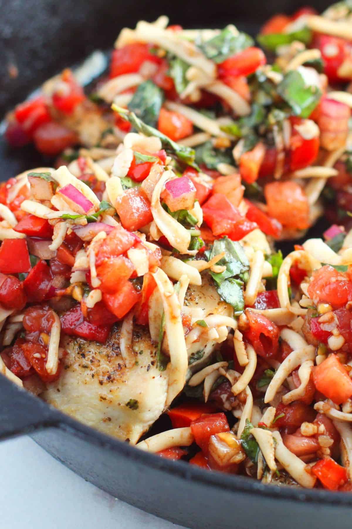 A close-up of a skillet with meat topped with a mixture of diced tomatoes, shredded cheese, red onions, and fresh basil.