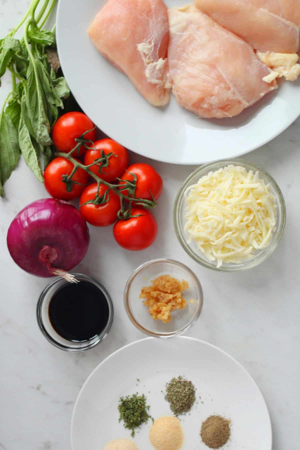 Raw chicken breasts, fresh basil, cherry tomatoes, shredded cheese, red onion, balsamic vinegar, minced garlic, and assorted dried spices are arranged on a white surface for a delicious bruschetta chicken recipe.