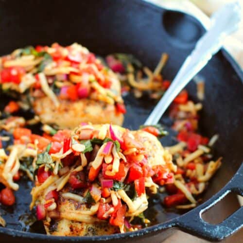Two cooked chicken breasts topped with diced vegetables and shredded cheese in a black cast iron skillet, inspired by a bruschetta chicken recipe, with a metal spoon resting inside.