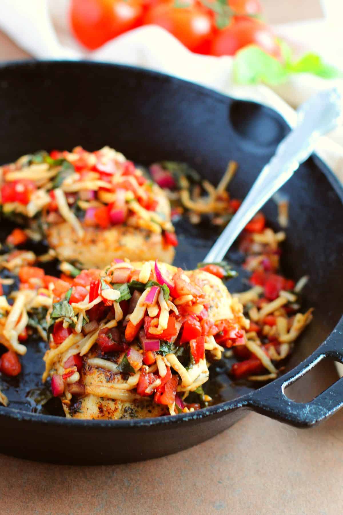 Two cooked chicken breasts topped with diced vegetables and shredded cheese in a black cast iron skillet, inspired by a bruschetta chicken recipe, with a metal spoon resting inside.