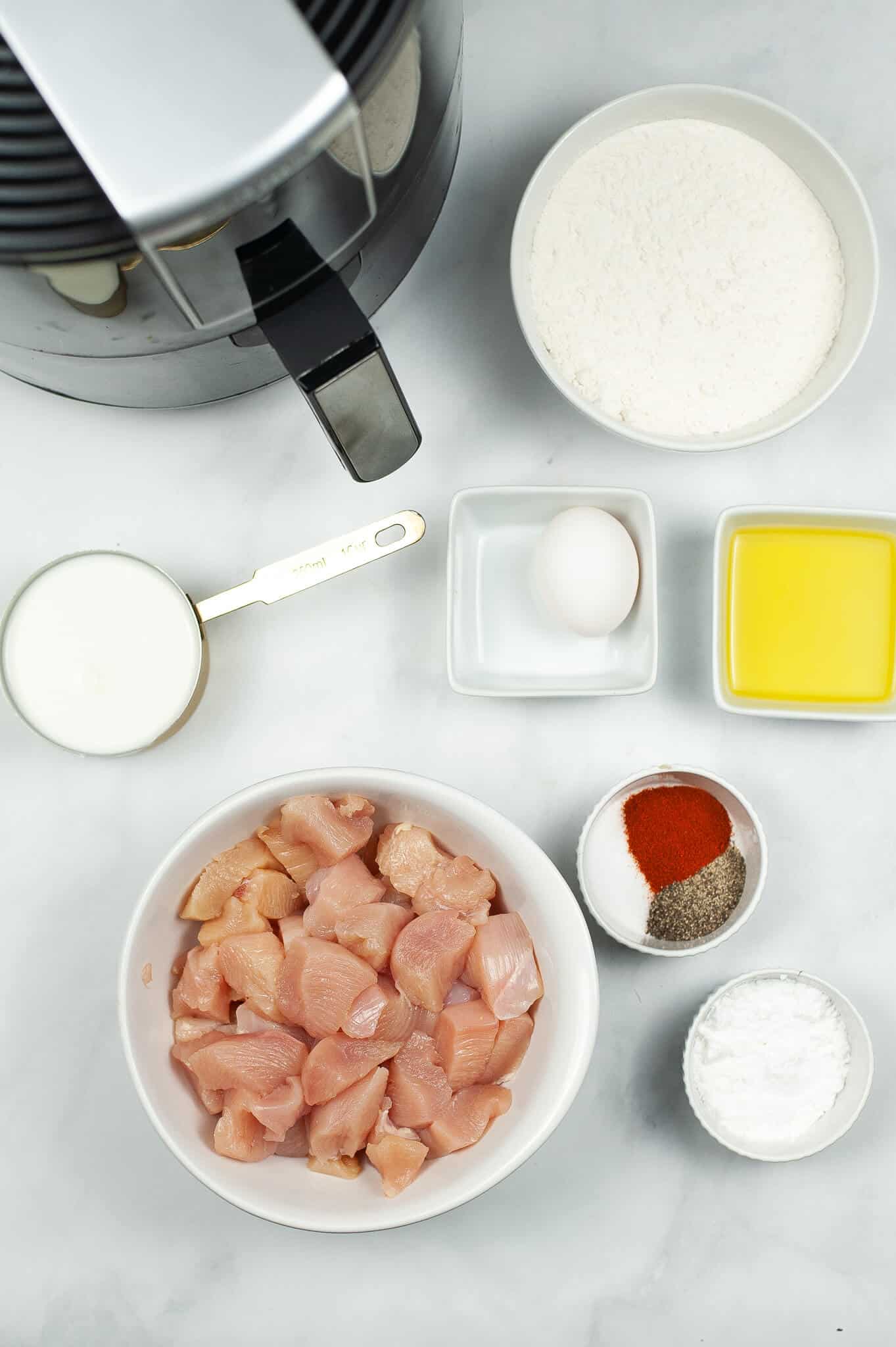 Top view of ingredients for copycat Chick-fil-A nuggets: an air fryer, a bowl of raw chicken pieces, flour, an egg, milk, melted butter, spices, and cornstarch arranged on a white surface.