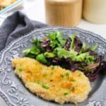 A Chicken Milanese cutlet garnished with herbs is served alongside a mixed green salad on a decorative gray plate.