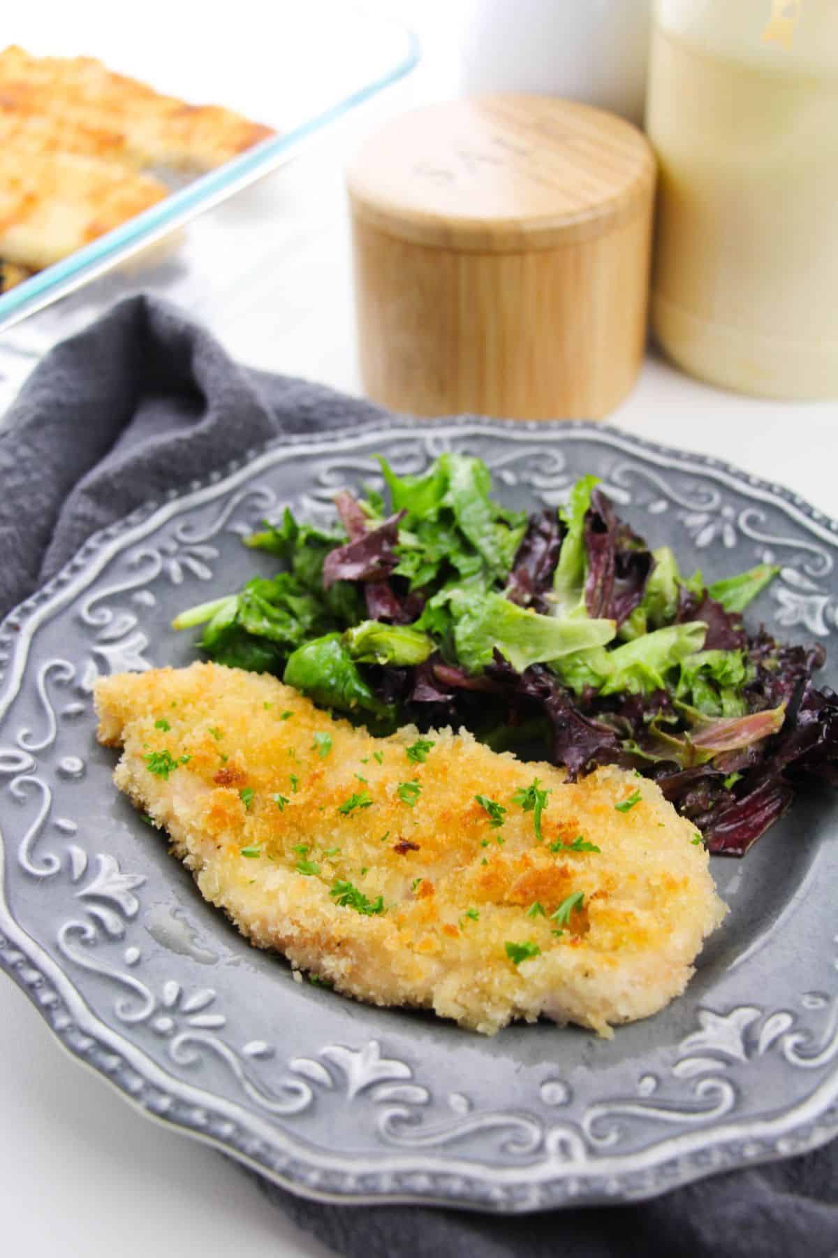 A Chicken Milanese cutlet garnished with herbs is served alongside a mixed green salad on a decorative gray plate.