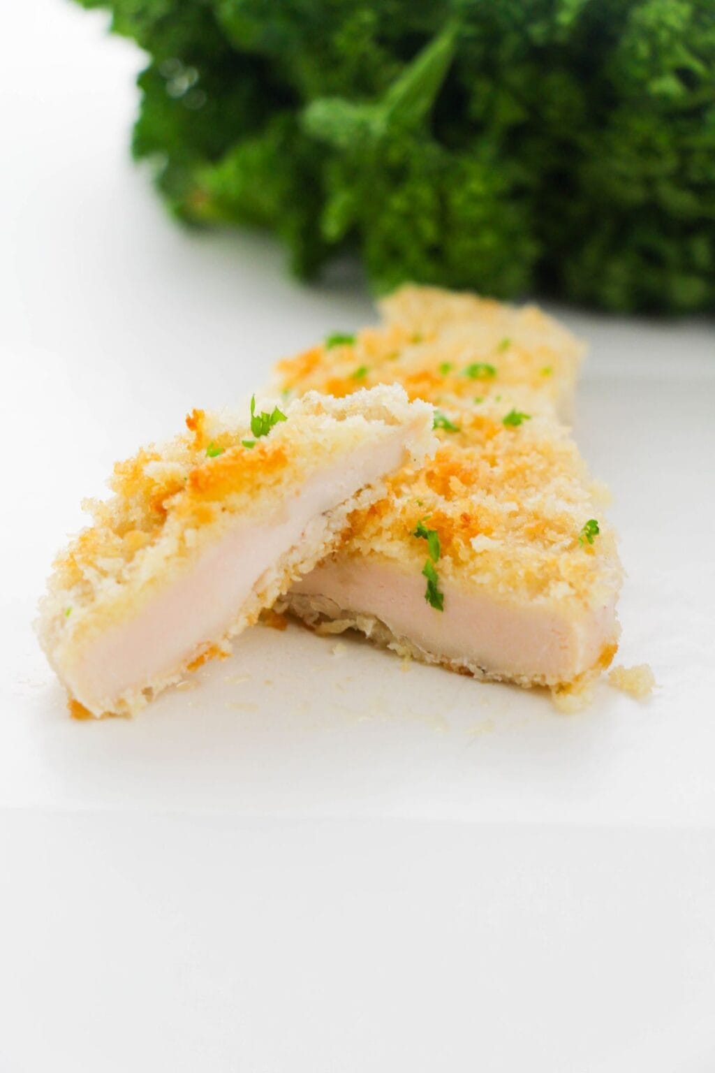 A breast, breaded and baked to perfection then sliced in half to reveal its juicy inside, with a bunch of leafy green parsley in the background.