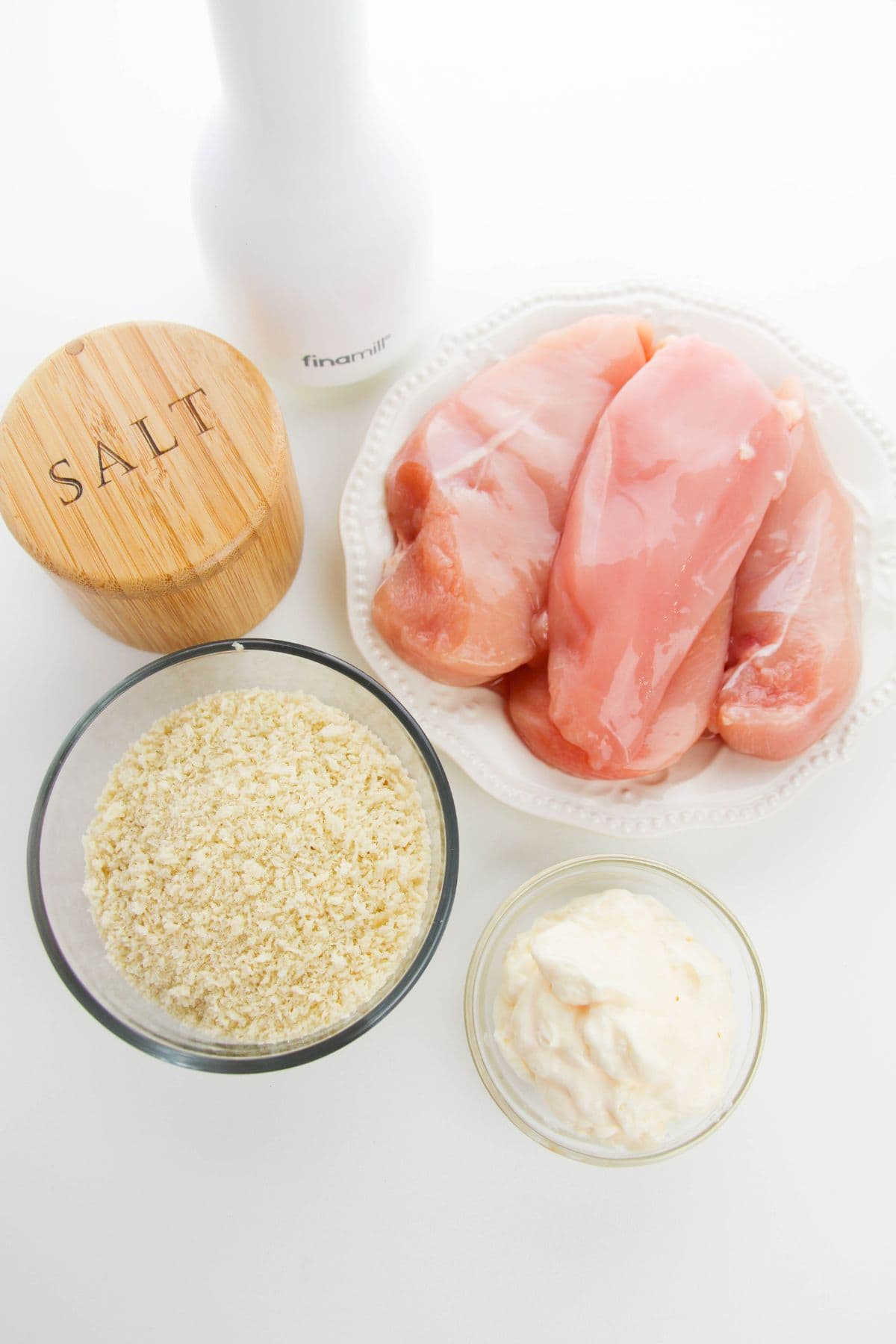 Raw chicken breasts on a plate, a small bowl of mayonnaise, a bowl of panko breadcrumbs, salt, and a white pepper grinder—a simple setup for preparing classic dish.