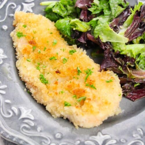 A classic Chicken Milanese—breaded, baked chicken cutlet—is served alongside a fresh mixed green salad on a decorative gray plate.