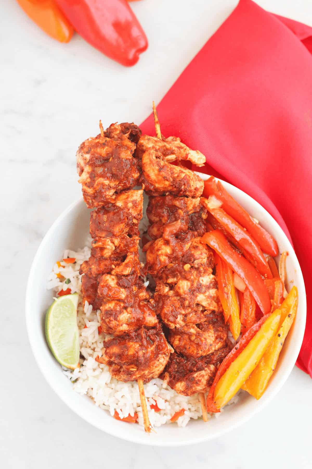 A white bowl filled with rice, meat, sliced grilled red and yellow bell peppers, and a lime wedge sits on a marble surface surrounded by fresh orange and red bell peppers and a red napkin.