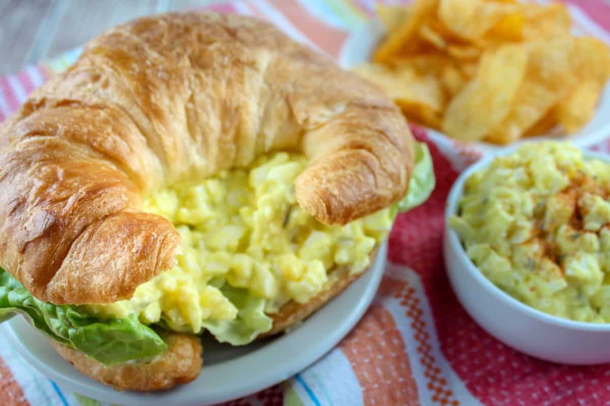 A croissant sandwich brimming with an easy egg salad recipe sits on a plate, accompanied by a bowl of potato chips and a side dish of the same delightful salad, all elegantly arranged on a colorful cloth.