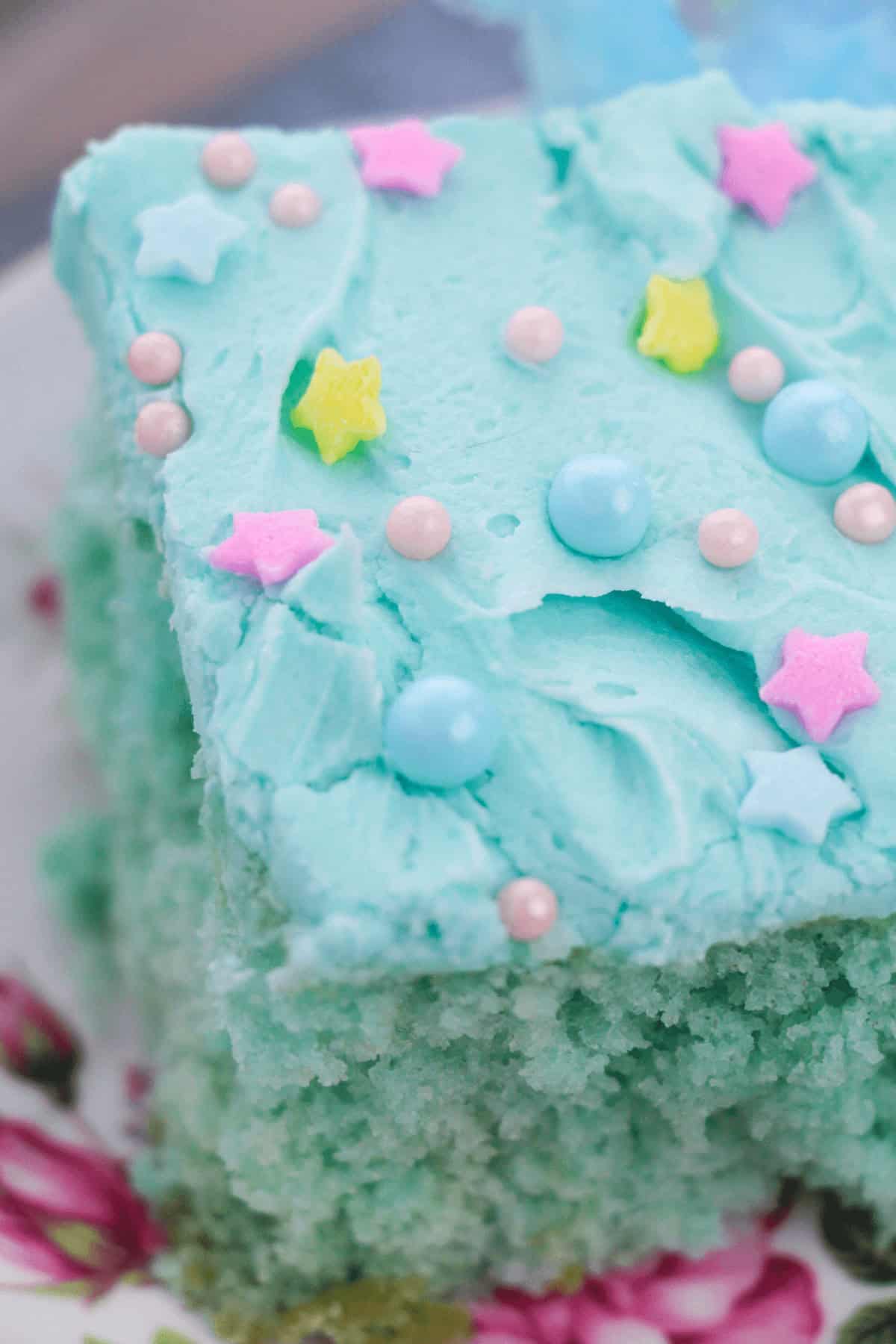 A slice of Cotton Candy Cake with blue frosting, adorned with pastel pink and blue round sprinkles and yellow and pink star sprinkles, rests elegantly on a floral plate.
