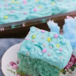 A slice of blue-frosted cotton candy cake with star and pearl sprinkles rests on a floral plate, while a tray of similarly delightful cakes lingers in the background.