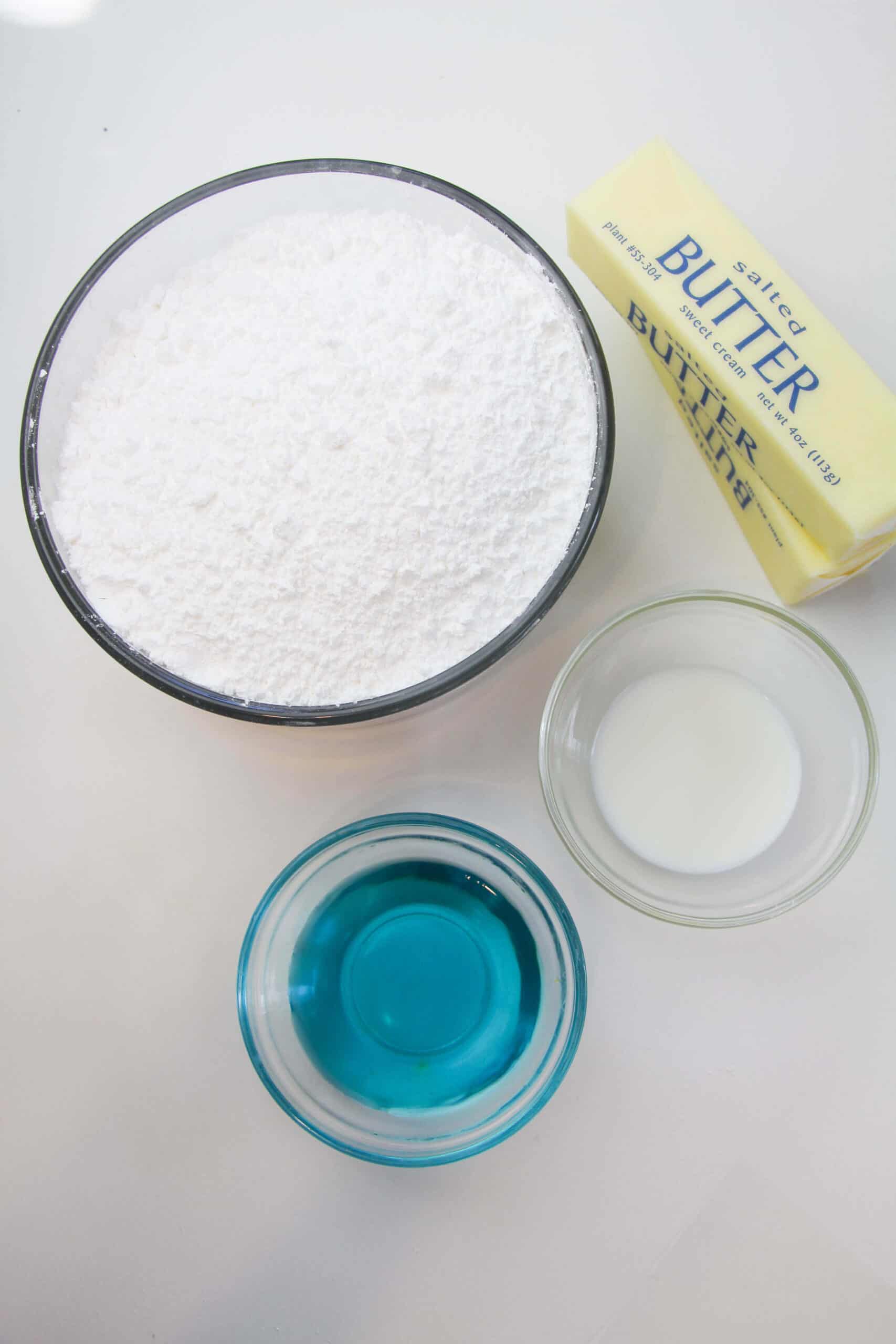 A bowl of powdered sugar, a stick of salted butter, a small bowl of milk, and a small bowl of blue liquid evoke the sweet allure all beautifully arranged on a white surface.
