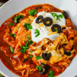 A bowl of Crockpot Chicken Enchilada Casserole-inspired soup topped with sour cream, shredded cheese, sliced black olives, and fresh cilantro.