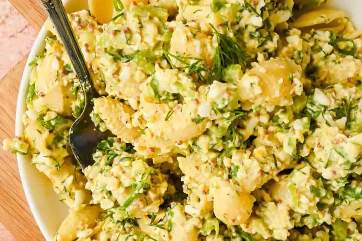 A bowl of deviled egg pasta salad with chopped herbs and a metal serving spoon.