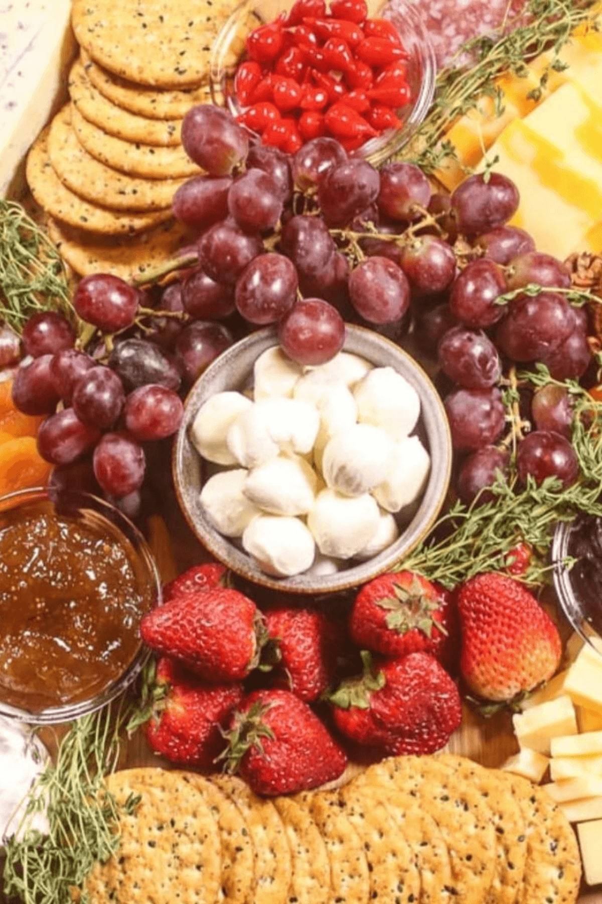 A spread of grapes, strawberries, mozzarella balls, crackers, and cherry tomatoes. Sprigs of herbs add a fresh touch, while a bowl of jam completes this simple yet elegant spread.