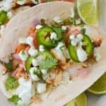 Two soft tacos filled with grilled chicken, diced tomatoes, chopped cilantro, sliced jalapeños, and creamy sauce—served alongside Fish Tacos with Cod and lime wedges on a white plate.