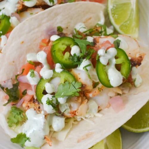 Two soft tacos filled with grilled chicken, diced tomatoes, chopped cilantro, sliced jalapeños, and creamy sauce—served alongside Fish Tacos with Cod and lime wedges on a white plate.