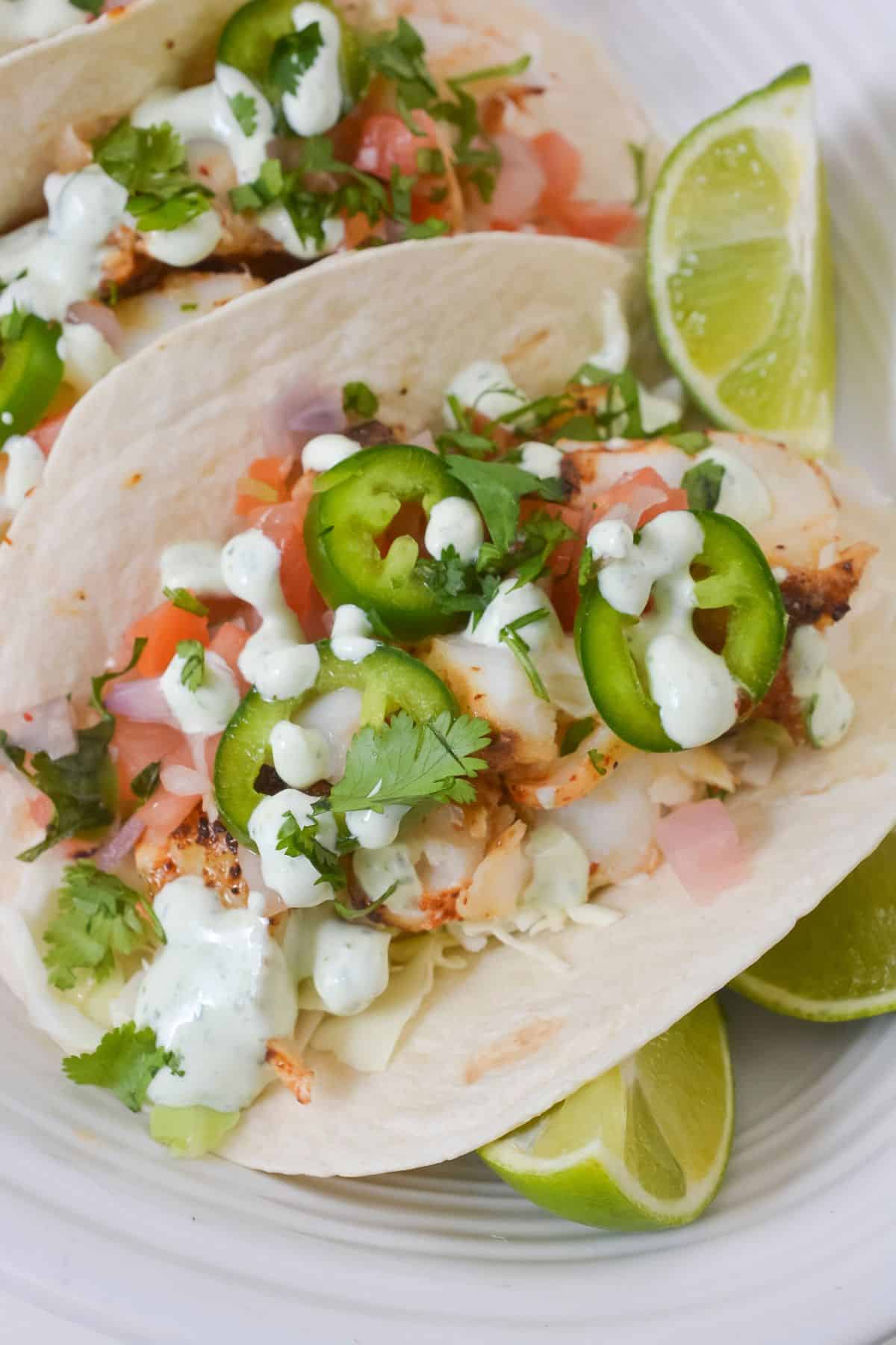 Two soft tacos filled with grilled chicken, diced tomatoes, chopped cilantro, sliced jalapeños, and creamy sauce—served alongside Fish Tacos with Cod and lime wedges on a white plate.