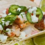 A close-up of Fish Tacos with Cod topped with sliced jalapeños, cilantro, diced tomatoes, and creamy sauce, served with lime wedges.