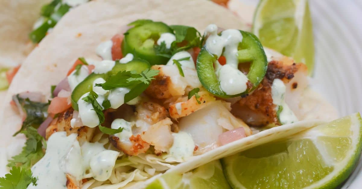 A close-up of Fish Tacos with Cod topped with sliced jalapeños, cilantro, diced tomatoes, and creamy sauce, served with lime wedges.