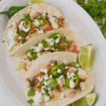 Three soft shell Fish Tacos with Cod filled with diced vegetables, herbs, and sliced jalapeños, topped with a drizzle of white sauce, served with lime wedges on a white oval plate.