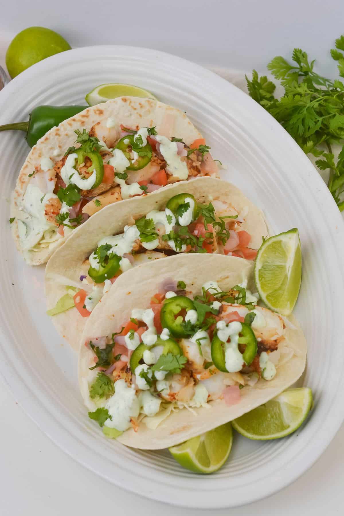 Three soft shell Fish Tacos with Cod filled with diced vegetables, herbs, and sliced jalapeños, topped with a drizzle of white sauce, served with lime wedges on a white oval plate.