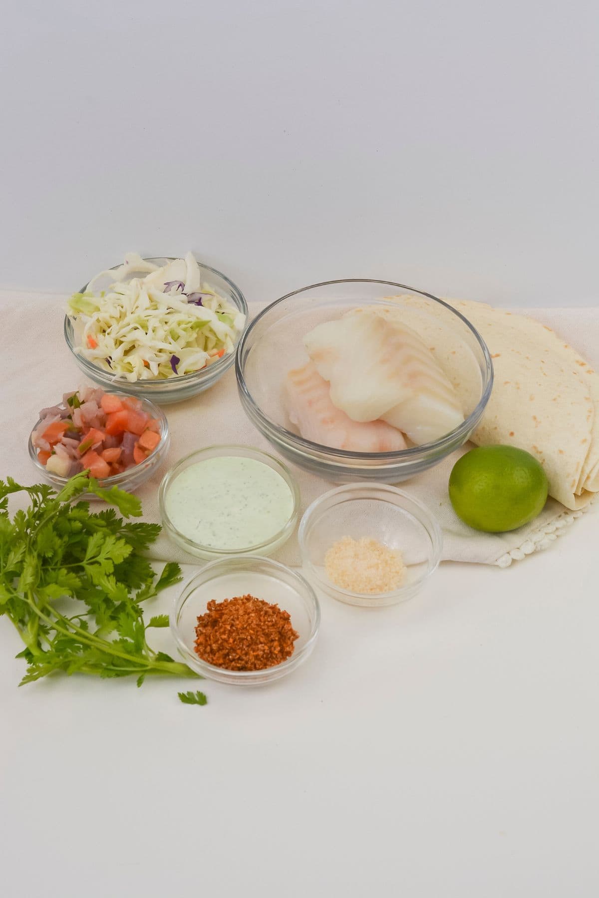 A variety of ingredients fillets, tortillas, shredded cabbage, pico de gallo, cilantro, lime, creamy sauce, grated cheese, and a small bowl of spices on a white surface.