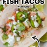 Close-up of Fish Tacos with Cod, topped with sliced jalapeños, cilantro, diced tomatoes, creamy sauce, and lime wedges on a plate. Text reads "Easy Fish Tacos with Cod—Fresh.