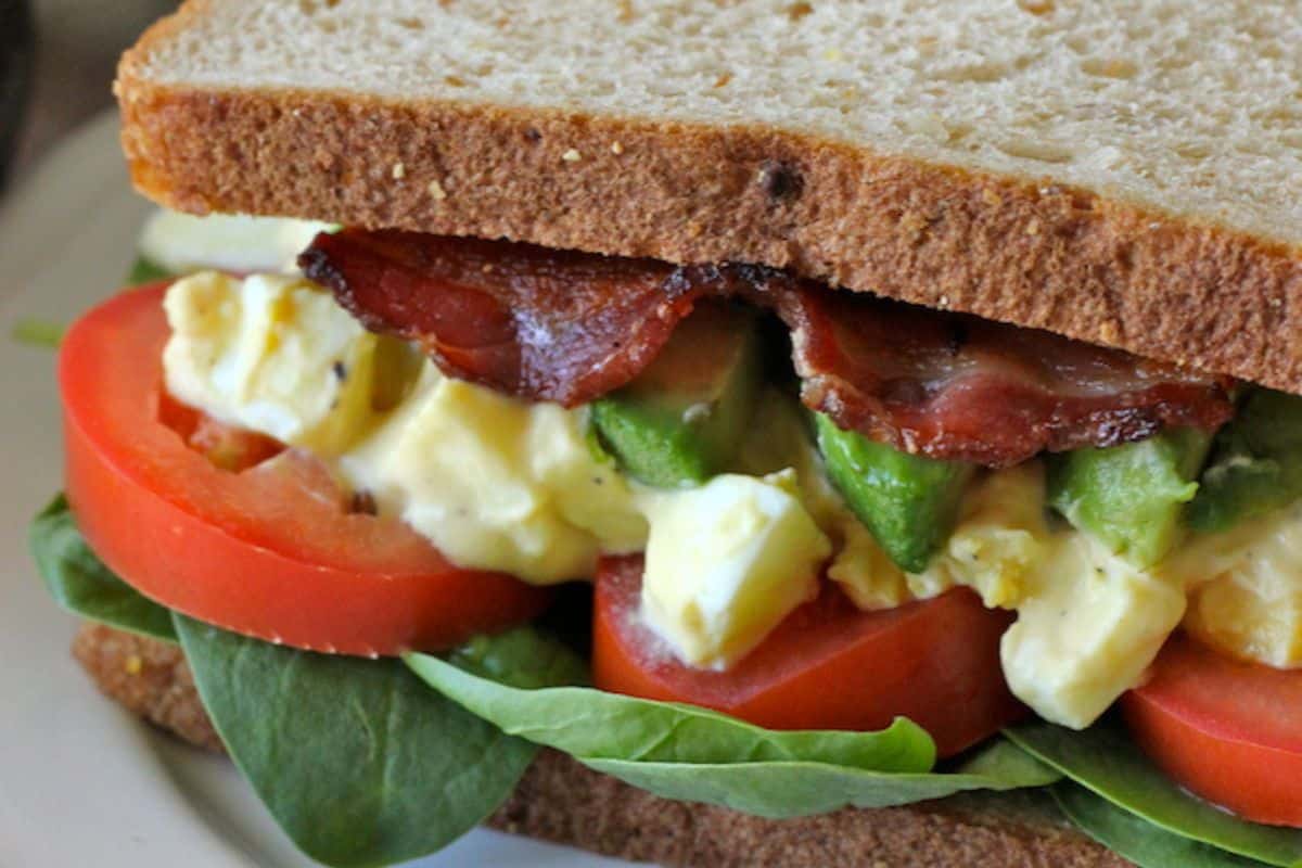 An easy egg salad recipe comes to life with avocado, bacon, fresh tomato slices, and spinach nestled between slices of brown bread.