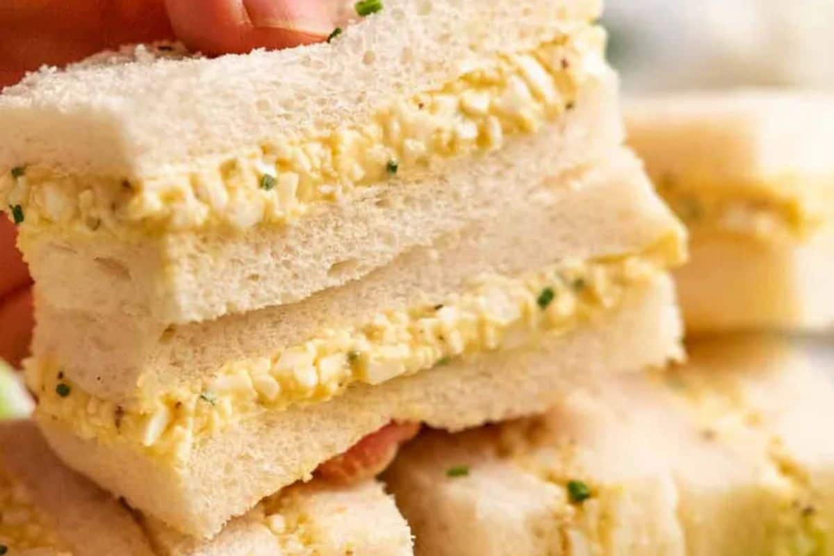 Three stacked egg salad sandwiches on white bread, featuring an easy egg salad recipe with a creamy filling and small chunks of egg, garnished with fresh herbs.
