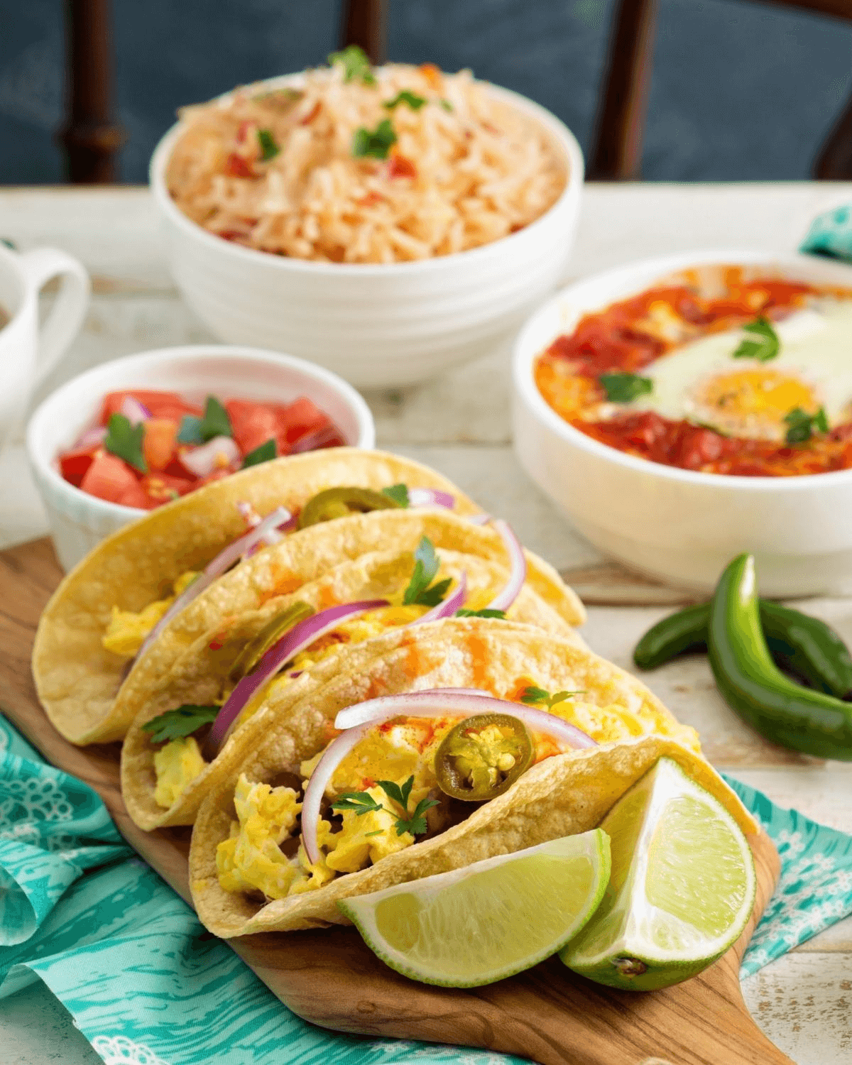 Three breakfast tacos with scrambled eggs, sausage, and avocado slices, garnished with lime wedges, served next to bowls of salsa and cheesy dip.