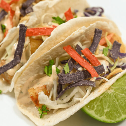 Two baja fish tacos with cabbage slaw and tortilla strips on a plate accompanied by lime wedges.