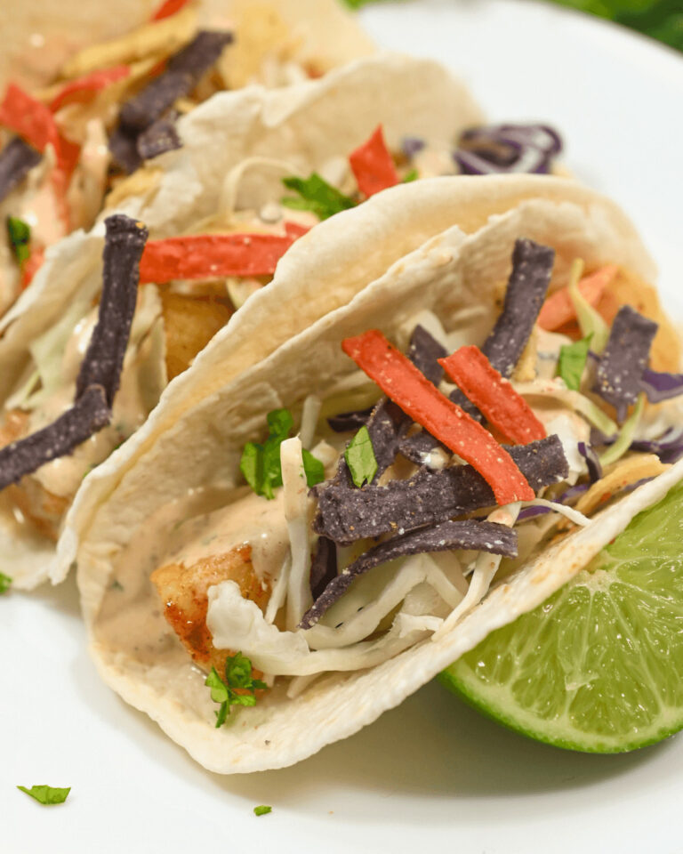Two baja fish tacos with cabbage slaw and tortilla strips on a plate accompanied by lime wedges.