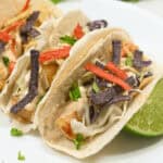 Two baja fish tacos garnished with purple cabbage, red pepper strips, and lime on a white plate.