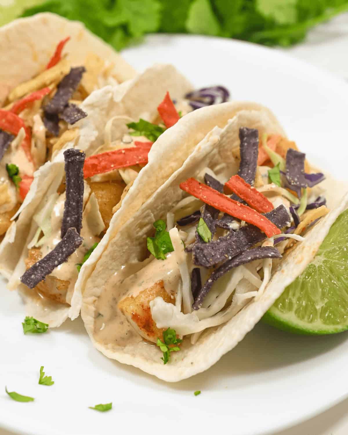Two baja fish tacos garnished with purple cabbage, red pepper strips, and lime on a white plate.
