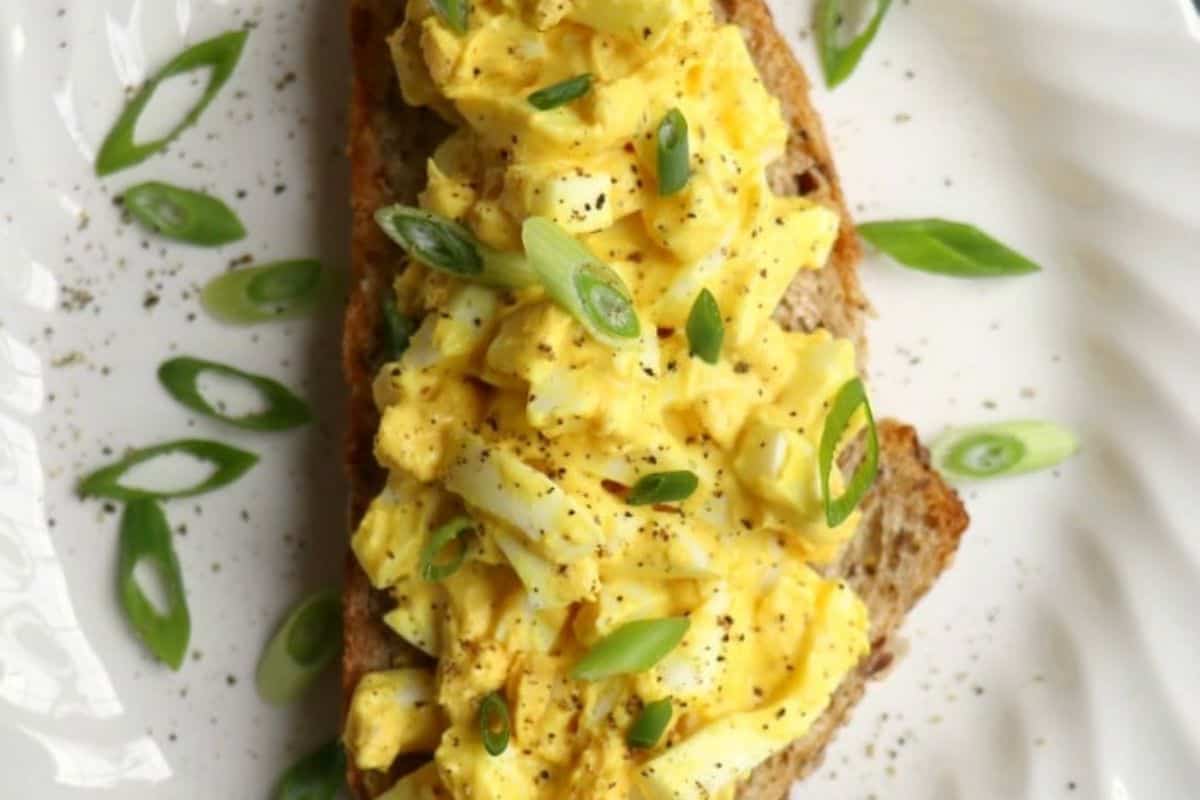 Easy egg salad recipes come to life with this delightful dish—egg salad with sliced green onions on a piece of bread, garnished with extra green onion rings and a sprinkle of black pepper, all beautifully served on a white plate.