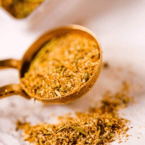 A measuring spoon filled with a rich blend of homemade chili seasoning rests on a marble surface, surrounded by a scattering of vibrant spices and herbs.