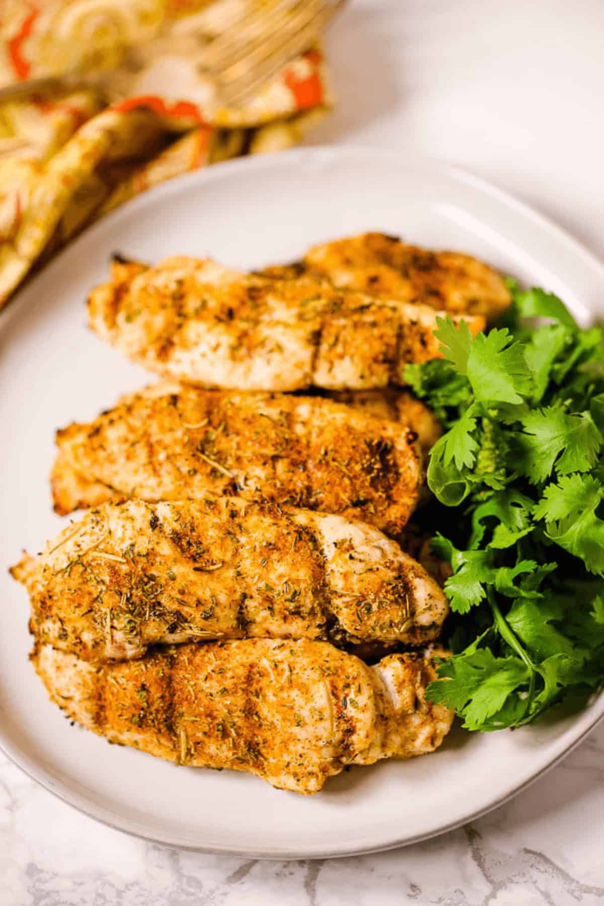 Grilled chicken breasts seasoned with a homemade chili seasoning and herbs on a white plate, garnished with a sprig of fresh cilantro.