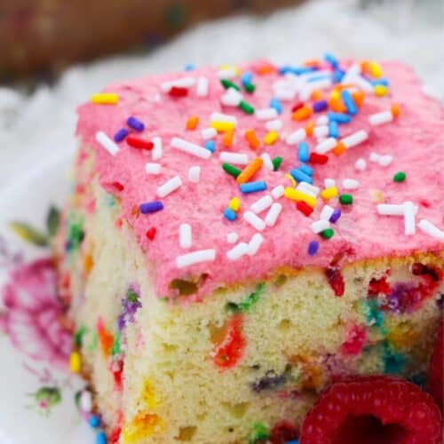 A vibrant slice of confetti cake with pink frosting and rainbow sprinkles rests elegantly on a floral plate, garnished with raspberries.