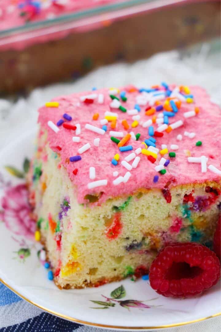 A vibrant slice of confetti cake with pink frosting and rainbow sprinkles rests elegantly on a floral plate, garnished with raspberries.