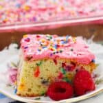 A slice with pink frosting and vibrant sprinkles, served on a plate alongside a few fresh raspberries.