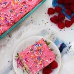A slice of confetti cake with pink frosting and colorful sprinkles on a floral plate, next to a basket of raspberries. The cake sits elegantly in a glass dish.