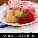 A delightful slice of confetti cake with pink frosting and sprinkles sits on a plate, elegantly garnished with fresh raspberries. In the background, a cake pan hints at more deliciousness to come.