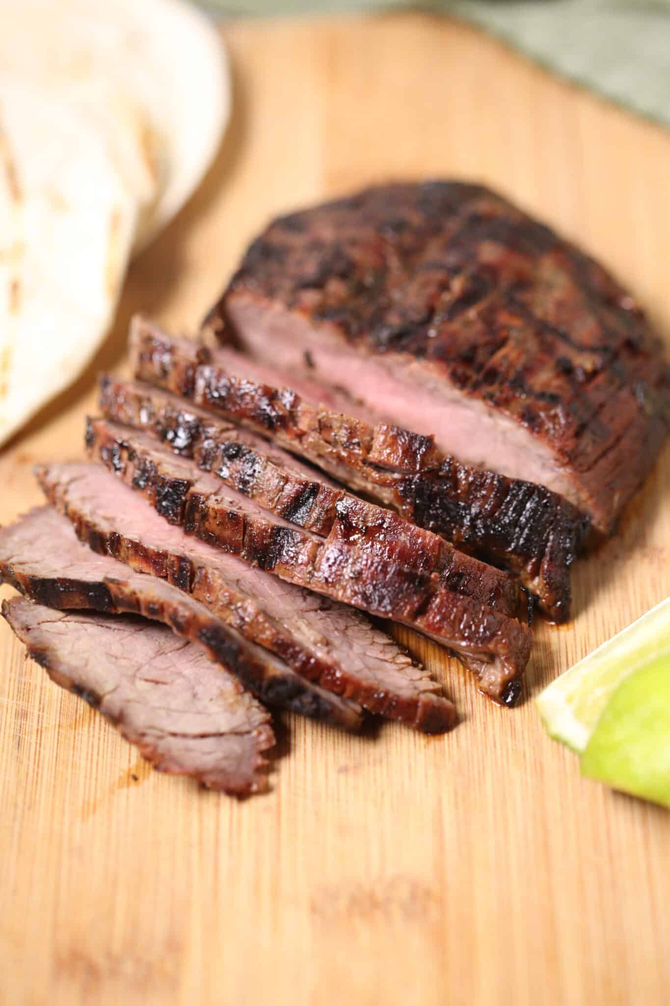 Sliced grilled steak for flavorful Street Tacos rests on a wooden board, with a lime wedge and tortillas ready in the background.