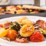 A plate of roasted Italian Sausage with Gnocchi, cherry tomatoes, broccoli, and shredded cheese sits in front, with a sheet pan of the same savory dish in the background.