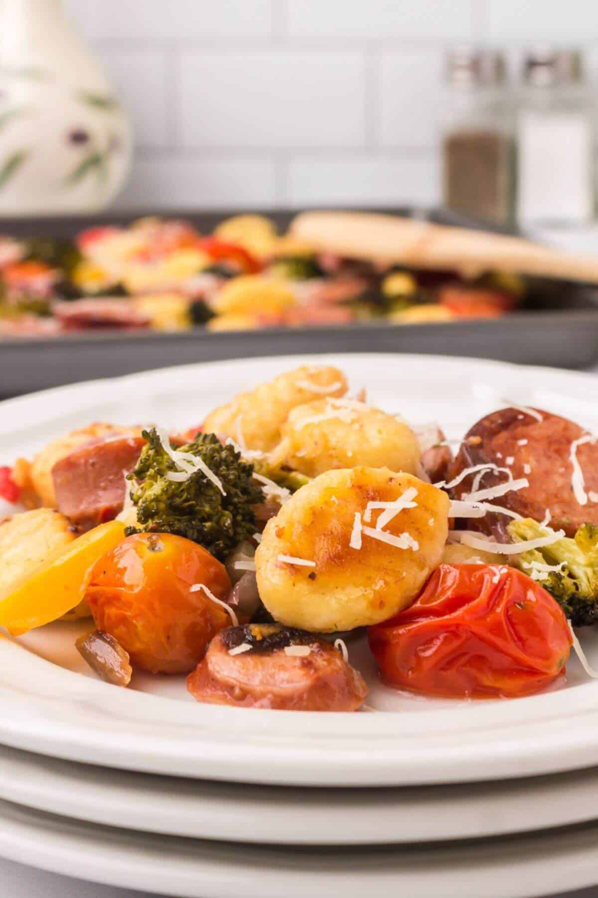 A plate of roasted Italian Sausage with Gnocchi, cherry tomatoes, broccoli, and shredded cheese sits in front, with a sheet pan of the same savory dish in the background.