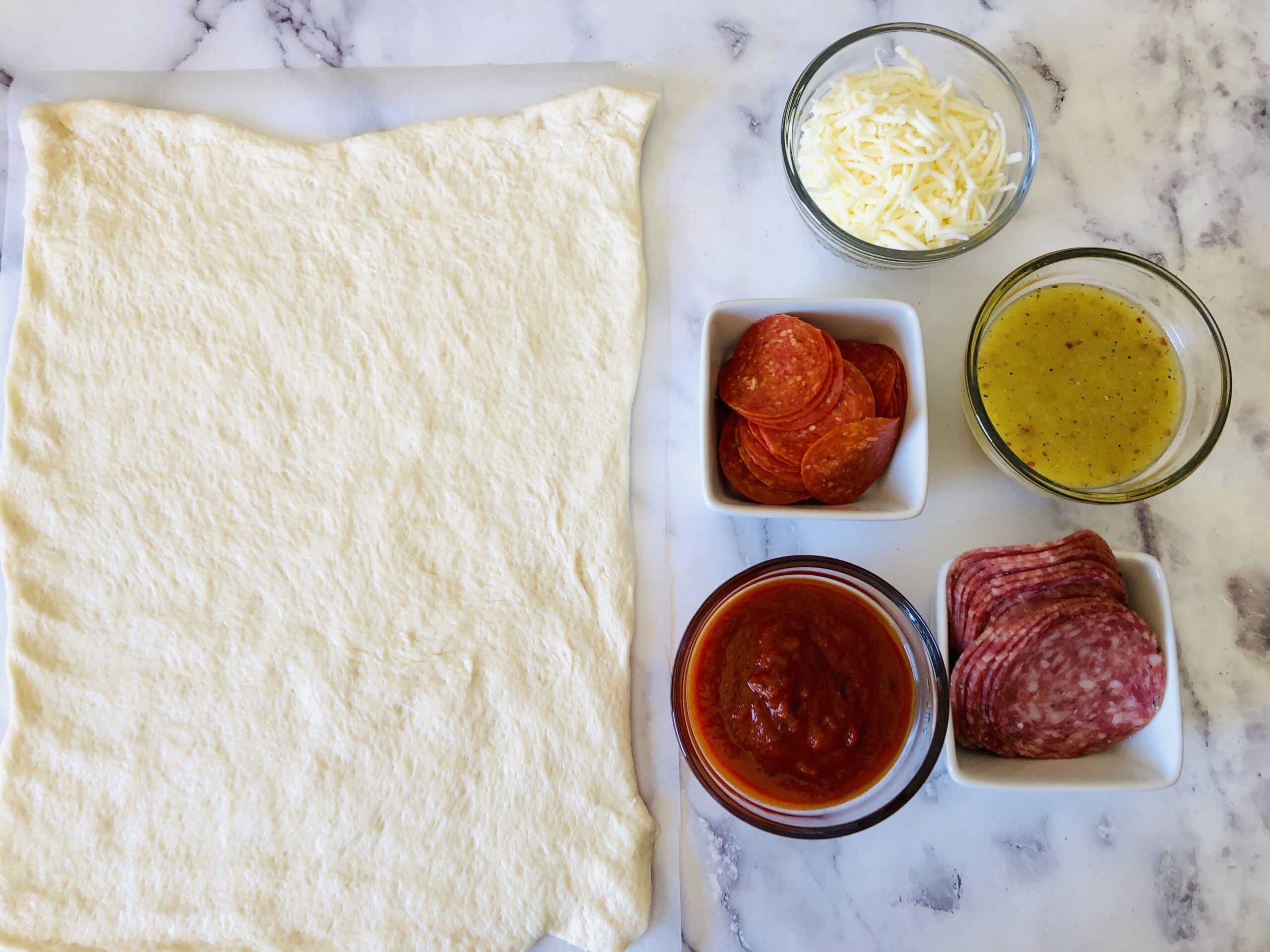 Ingredients for making Italian Stromboli displayed on a marble countertop, including dough, shredded cheese, pepperoni slices, salami, tomato sauce, and herb-infused oil.