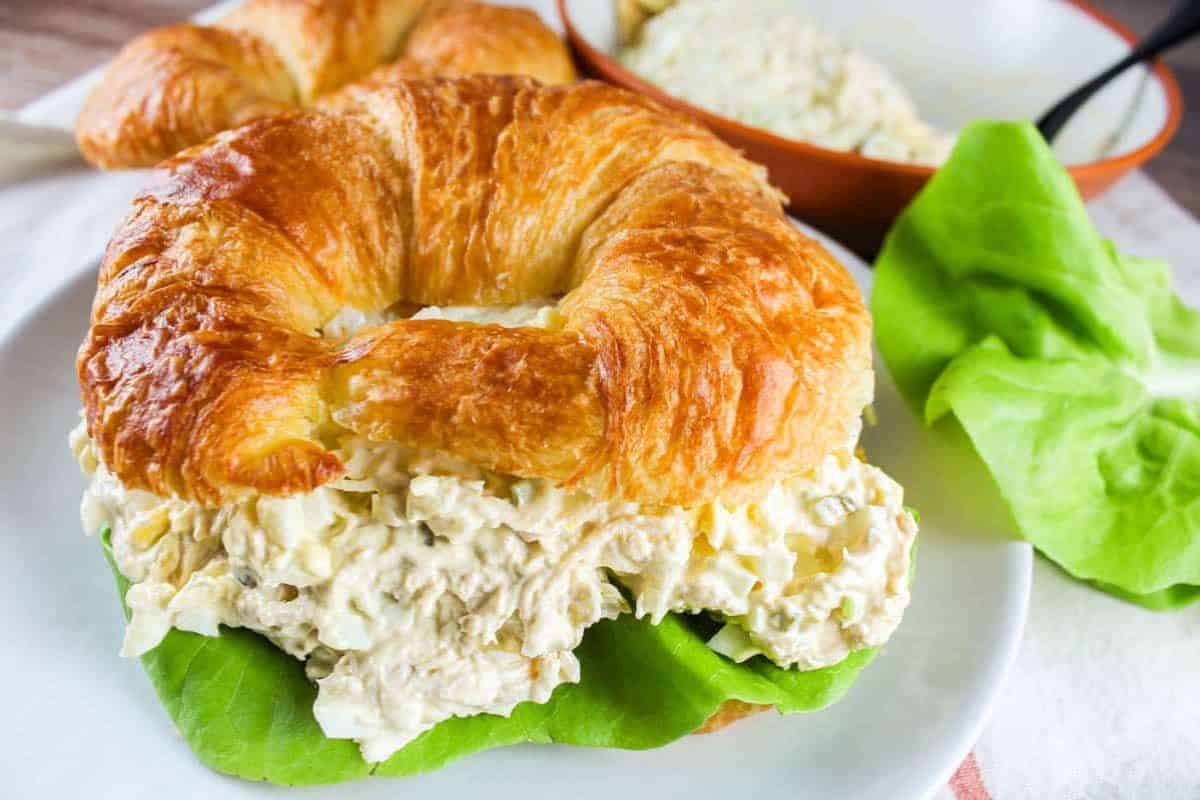 A croissant sandwich, brimming with creamy egg and chicken salad, rests on a plate adorned with fresh lettuce. In the background, a bowl offers more filling inspiration for easy egg salad recipes.