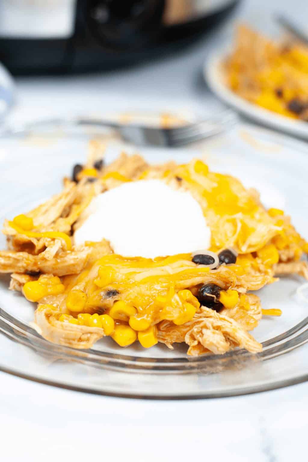 A glass plate showcasing a vibrant Slow Cooker Mexican Chicken Lasagna features layers of shredded chicken, corn, black beans, and melted cheese, topped with a dollop of sour cream. A fork awaits in the background.