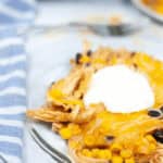 A clear glass dish showcases layers of Slow Cooker Mexican Chicken Lasagna, with shredded chicken, corn, black beans, melted cheese, and a dollop of sour cream. A blue striped towel is beside it.